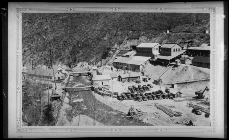 Image of a photograph overlooking an unidentified mining complex. Several buildings, vehicles, and a river are visible.