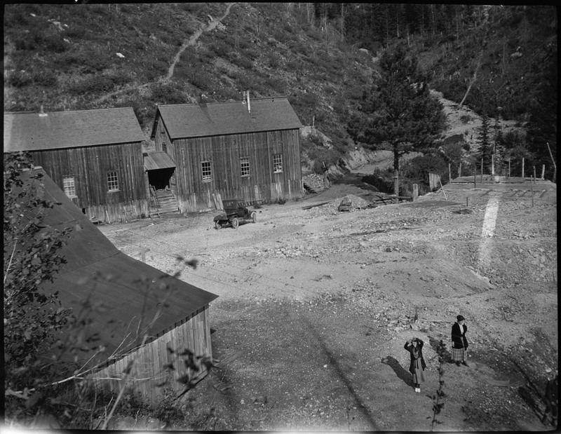 Two women standing outside of a mine. The photo is taken from an elevation and one woman is looking up at the photographer. There are buildings and a car in the background.
