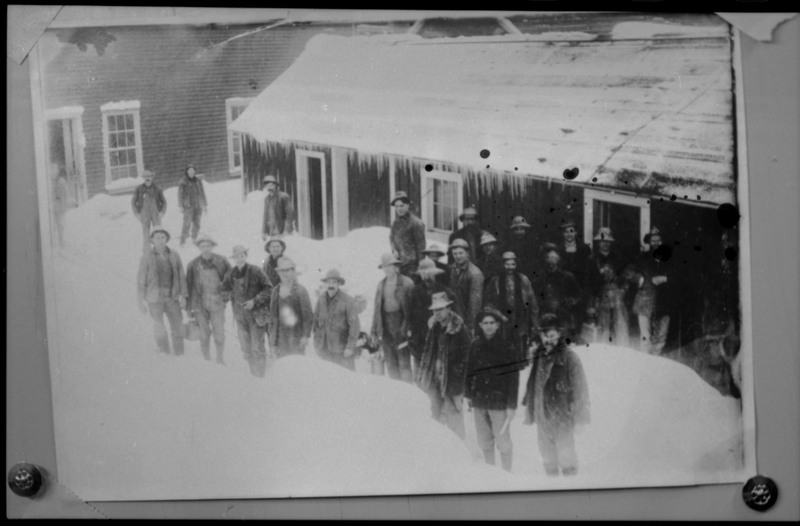 Image of a photograph of a group of miners standing outside in the snow. There are a couple buildings behind them. Unclear what mine they work at.