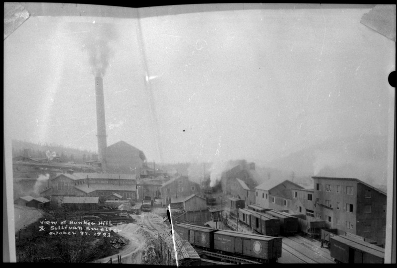 Image of a photograph of the view of Bunker Hill and Sullivan Smelter. Several buildings are visible, as well as train cars.