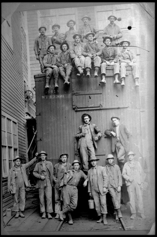Twenty men standing on or near a train car. Eleven of the men are on top of the roof of the train car while the rest are on the ground. They are all looking at the photographer.