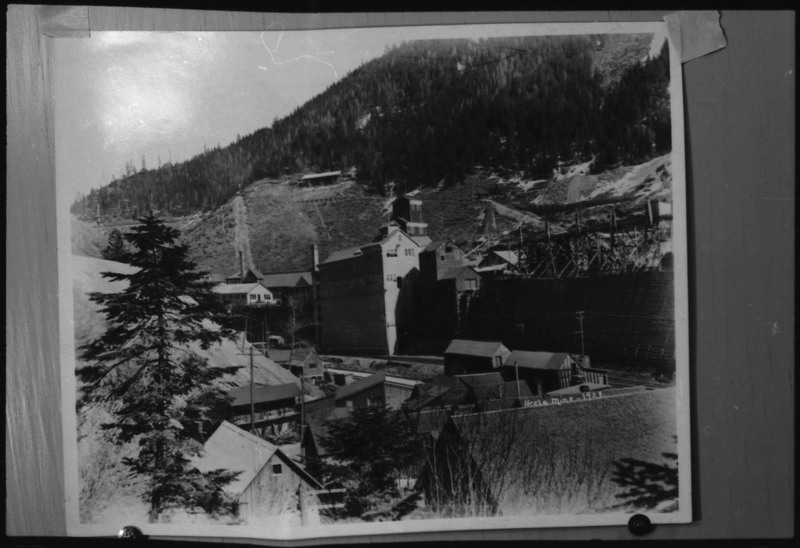 Image of a photograph of Hecla Mine in Burke, Idaho. Some of the buildings of the complex are visible, as is the tree line on the hills surrounding the mine.