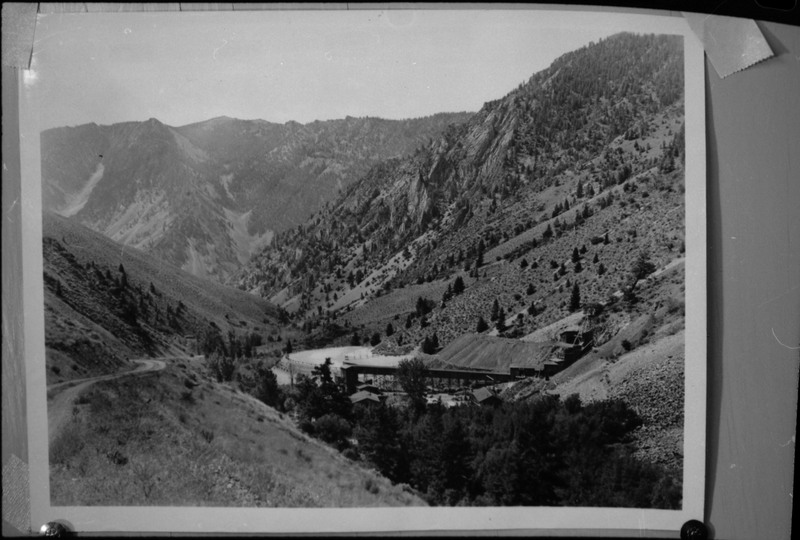 Image of a photograph that overlooks an unidentified mine. Most of the buildings are obscured by trees, but the mine entrance is visible, as is the mountains in the background.
