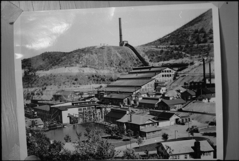 Image of a photograph of an unidentified mining complex. Several buildings are visible, and there appears to be a smelter on the top of a hill behind the complex.