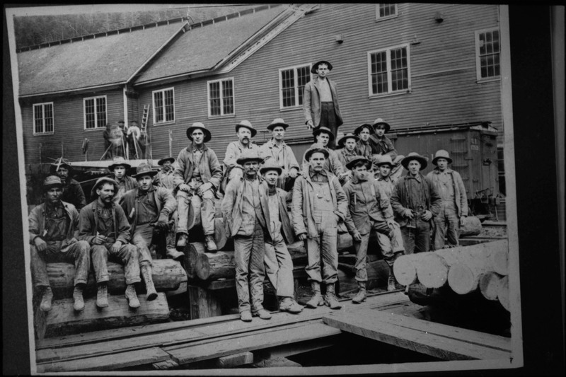 Image of a photograph of a group of unidentified men. They are standing and sitting on top of stacked wooden logs, posing for a photo in front of a building. They are likely miners or mill workers.