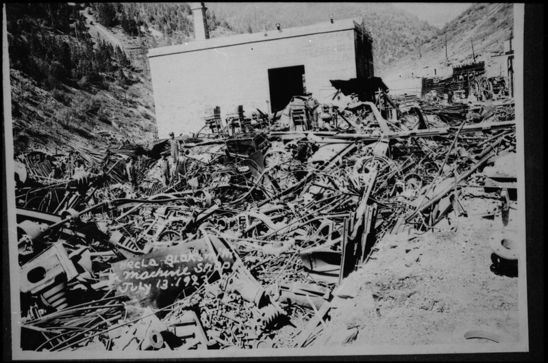 Image of a photograph of the Hecla blacksmith and machine shop in Burke, Idaho. The shop building is in the background of the photo behind a very large pile of scrap metal.
