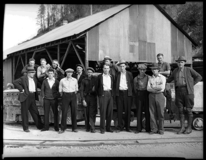 A group of men standing outside of Silver Summit Mine. They are all well dressed and standing in front of a building. Some men are standing on minecarts with most men standing in front of it.
