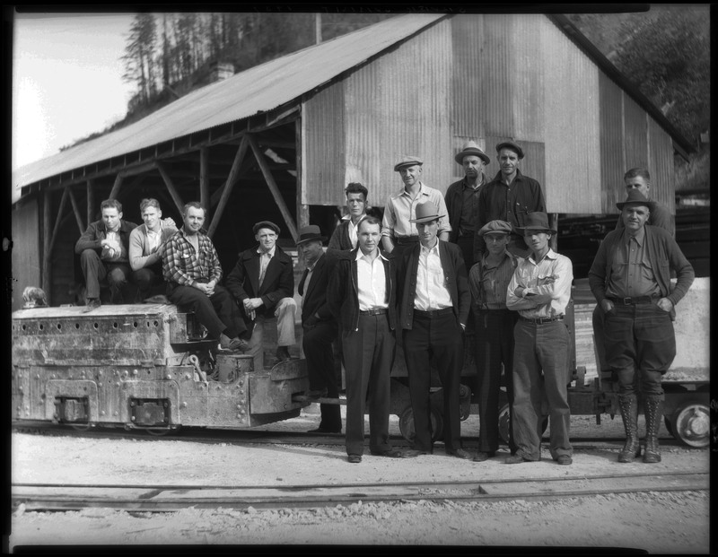 A group of men standing outside of Silver Summit Mine. They are all well dressed and standing in front of a building. Some men are standing on minecarts with most men standing in front of it.