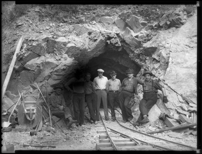 Seven men at the entrance of an unidentified mine. They are all sitting or standing in the mine entrance over the mine cart tracks, and they are all looking at the camera. One man has his face smudged out.