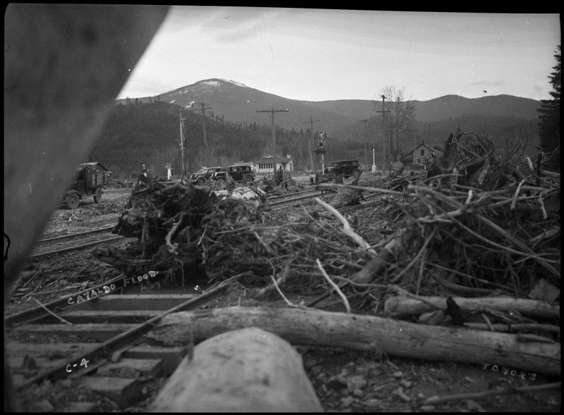 Downed trees and other plant debris litter a train track after the Wallace flood. Several buildings and telephone poles are visible in the background, as well as a sign that says "Place No Cars This Point [unintelligible]." Frost Peak is also visible in the far background.