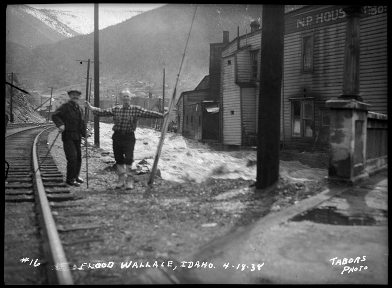 The Wallace, Idaho flood in front of some residential houses. There are two men standing between the water and some railroads.
