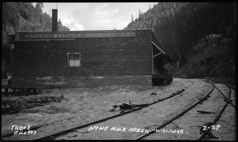 The flooding caused by Ninemile Creek in Wallace, Idaho. Water is rushing past a building with a sign that reads "Pacific Fruit and Produce Co."