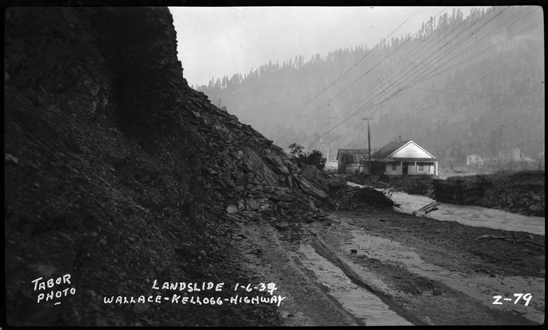 A landslide on the Wallace-Kellogg Highway near Wallace, Idaho. The landslide occurred over a dirt road directly next to a stream. A house and telephone pole are visible in the background.