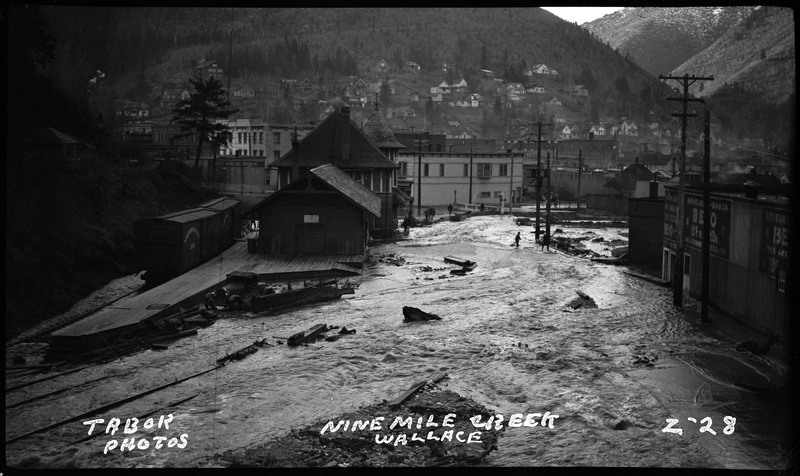 Ninemile Creek in Wallace, Idaho during a flood. The water is over the streets and rushing up to several buildings, and some damage can already be seen.