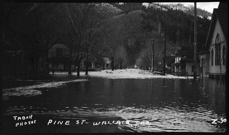 Pine Street in Wallace, Idaho during a flood. There is water overflowing in the streets and up to the houses and church.