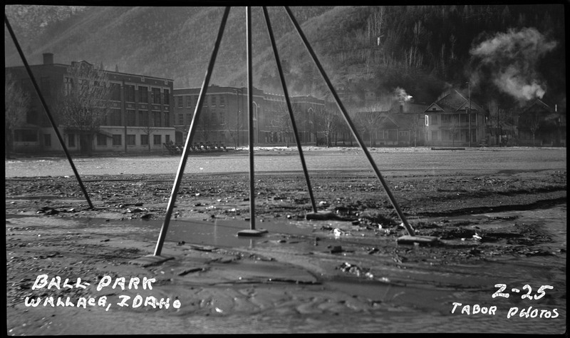 Image of the ballpark in Wallace, Idaho after flooding. Flooding appears to recede at the Wallace Ball Park during the Wallace flood. Much of the semi-dry land appears to be mud and rock. Buildings can be seen in the distance across from the ballpark.