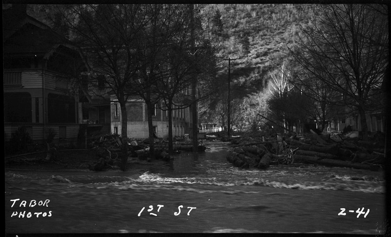 Street view of 1st street in Wallace, Idaho during the Placer Creek flood. Water is rushing in the street and fallen timber can be seen.