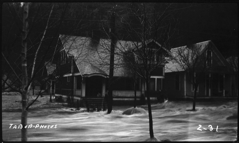 Image of a house behind some bare trees during the Placer Creek flood. Water is rushing down the street.