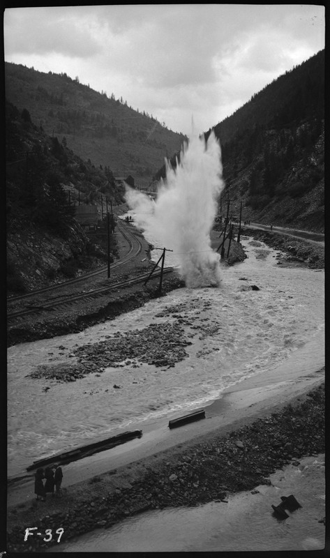 Image of what looks like an explosion near damaged railroad tracks during a flood. Three people can be seen watching the possible explosion on the bottom left.
