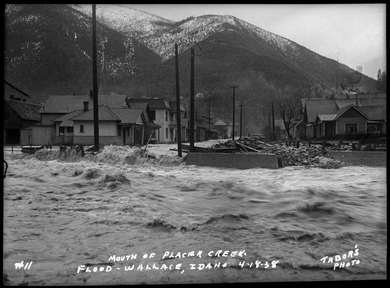 A view of the mouth of Placer Creek during the Wallace flood. There are several buildings, telephone poles, and piles of debris in the background.