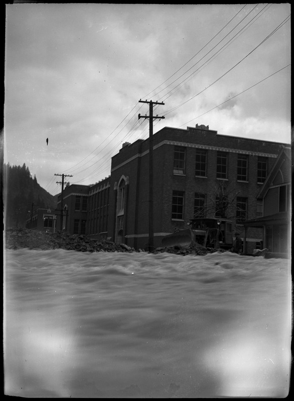 Rushing water in front of buildings and a man standing next to a bulldozer.