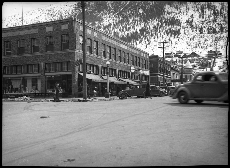 Street scene in Wallace, Idaho. Automobiles are parked along the road. A few people are walking on the sidewalk. A woman can be seen crossing the road. A snow covered mountainside with trees is in the background.