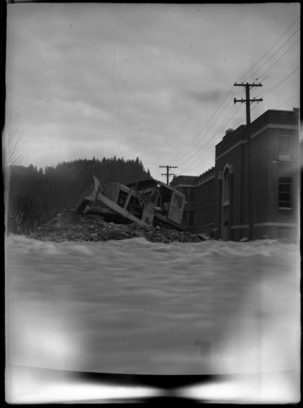 A vehicle is perched atop a pile of debris in the middle of the flood waters. Several buildings and telephone poles are visible in the background.