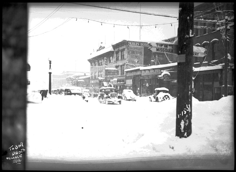 Snowy street scene. Automobiles are parked and in the street. A person can be seen on the left shoveling snow. A signpost on the right shows the direction to Missoula, Spokane, and other cities.