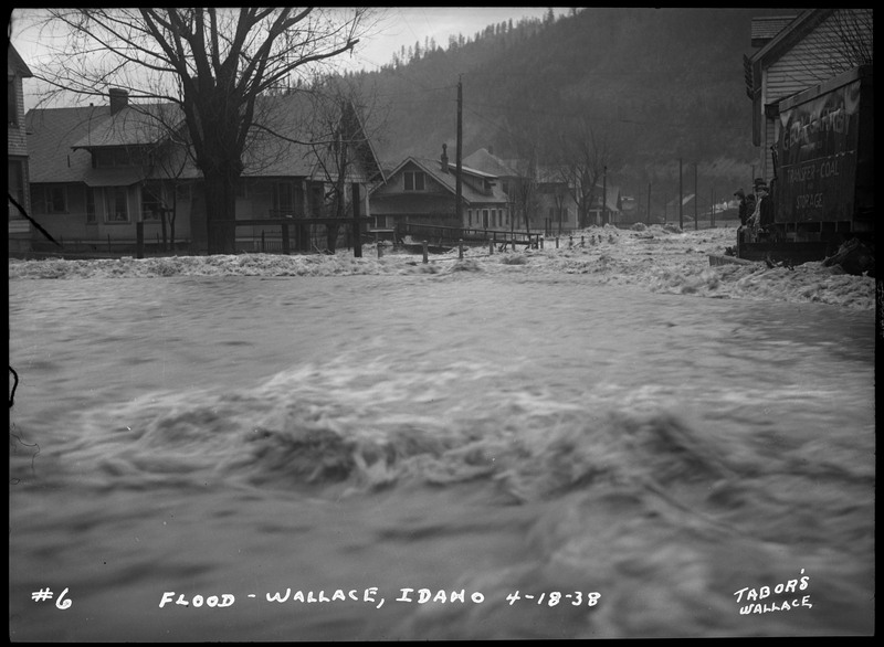 Water rushes near several buildings during the Wallace flood. There are also trees and telephone poles in the background.