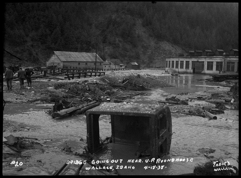Bridge going out near U.P. Roundhouse in Wallace, Idaho during a flood. Three people are standing on the left side of the image surveying the damage caused by the flood. Debris including a large machinery part is strewn about.
