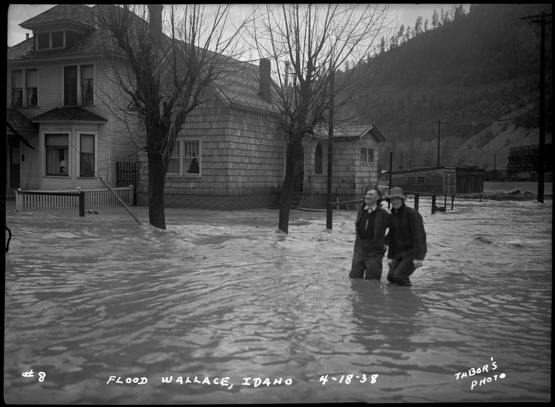 Two people wading through flood waters. The water has reached up to the houses nearby.