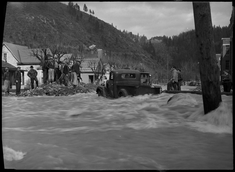 A large group of people watch as a person attempts to drive a truck through the flood waters. Several trees and houses are visible in the background.