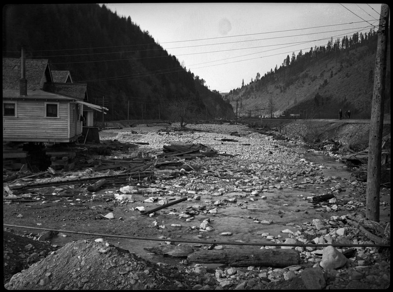 Damage and debris after a flood in Wallace, Idaho. A building is still standing on the left. Two men can be seen in the distance walking on the road on the right.