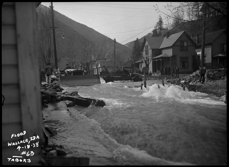 Water rushes through a neighborhood during a flood in Wallace, Idaho. A few people stand on nearby dry land.