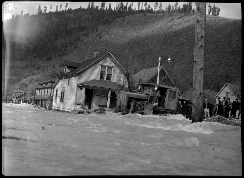 People gathering near a vehicle stuck in a flood in Wallace, Idaho as water rushes back. The people and vehicle are in front of a few standing houses.
