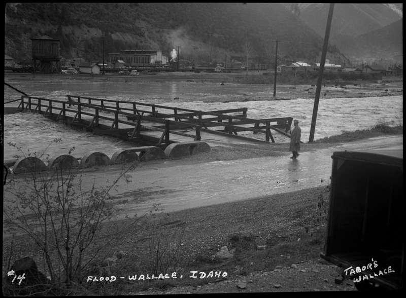 Water covers the surface of a bridge during the Wallace flood. A man standing on a road nearby appears to be looking at the flood waters. There are several buildings and telephone poles in the background.