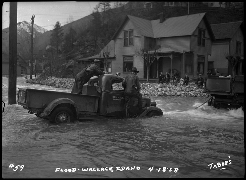 Two men can be seen in an automobile in the floodwater at the corner of River Street and 2nd Street. Several people watch while sitting on rocks.