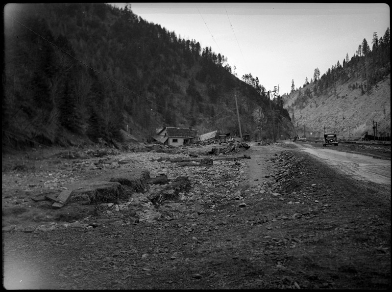 Landscape image depicting the damage caused by flooding. A few damaged houses are in the distance along with debris. A car is on the side of the road on the right.