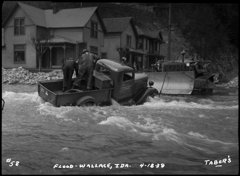 Two men can be seen in an automobile in the floodwater at the corner of River Street and 2nd Street. Several people watch while sitting on rocks. Another man is in a bulldozer.