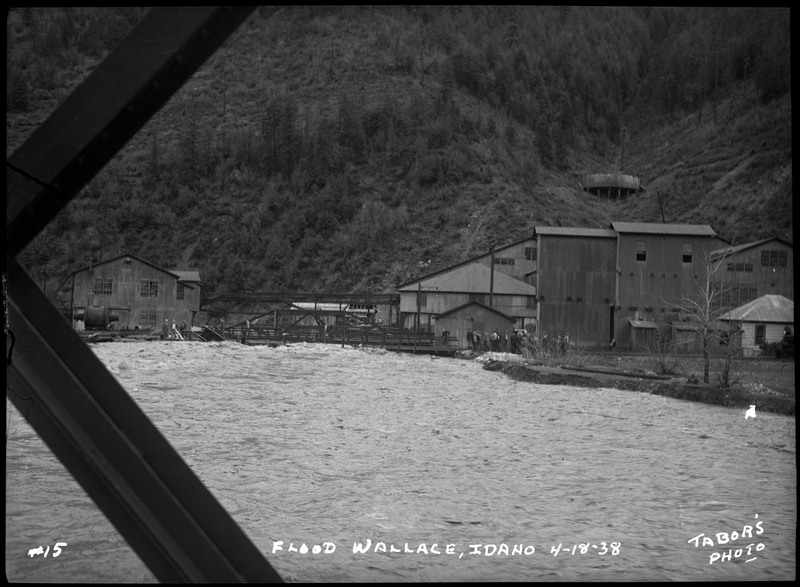 Water rushes near several buildings during the Wallace flood. It appears that the photo was taken from a bridge. A small crowd of people is nearby on dry land and a neighboring bridge.