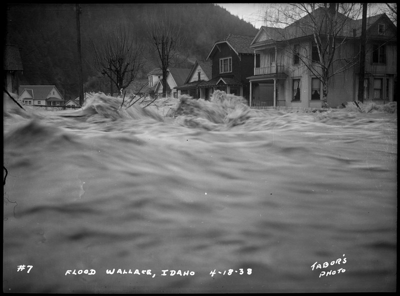 Water rushes near several buildings during the Wallace flood. There are several trees and telephone poles in the background. It appears the water reaches the bottom of first story windows.