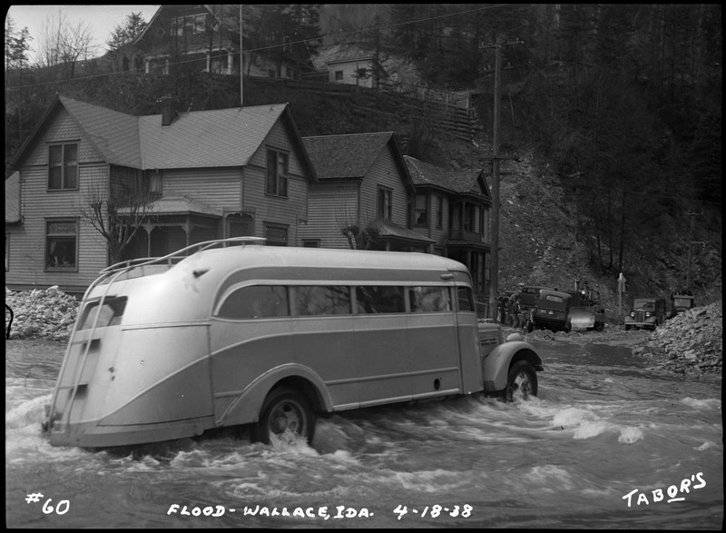 A car attempts to drive through flood waters in Wallace, Idaho. There are several houses, other cars, and unidentified people in the background.