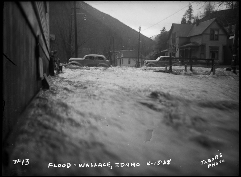 Water rushes past several buildings during the Wallace flood. Two cars appear to be driving through the flood waters.