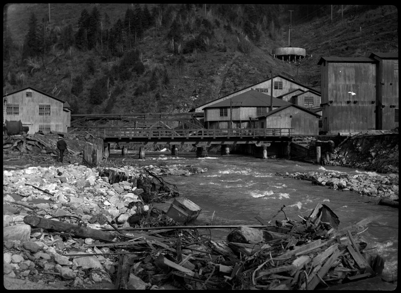 Image of some buildings and a bridge possibly during or after a flood in Wallace, Idaho. A man can be seen walking along the debris on the side of the water. Debris from the flood can be seen in the foreground.