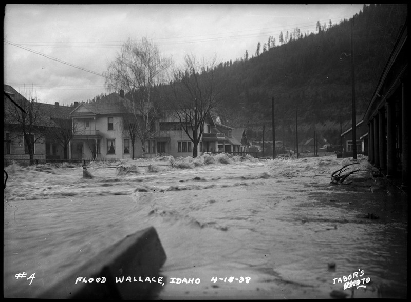 Image of a rushing water covering the street during a flood in Wallace, Idaho. There are buildings, trees, and telephone poles still standing.
