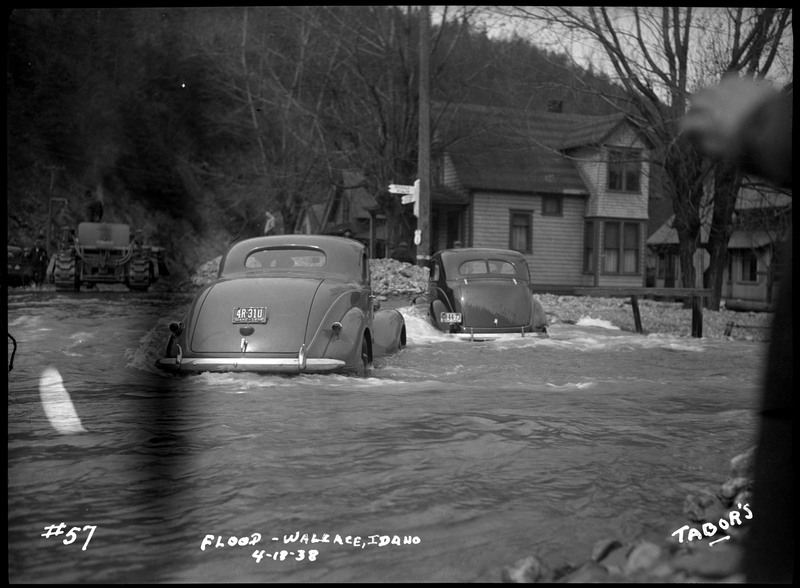 Two automobiles in the water during a flood in Wallace, Idaho. A few buildings and a tractor are in the background. Part of a hand can be seen on the right.