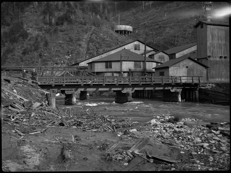 Image of some buildings and a bridge possibly during or after a flood in Wallace, Idaho. Two men are standing on the left end of the bridge in the distance. Debris from the flood can be seen in the foreground.