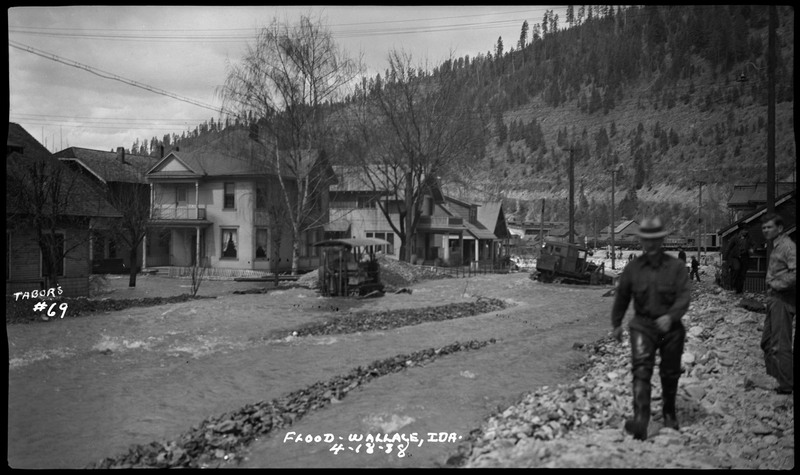 Street view of Wallace, Idaho during or after a flood. People can be seen in the distance, walking or standing. A man wearing boots and a hat is walking towards the camera. Two tractors or pieces of machinery are stuck in the street.