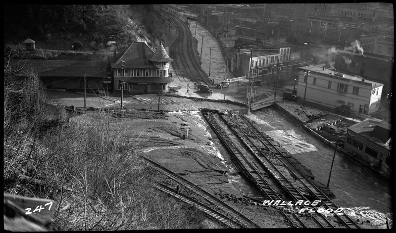 A view from above of the damage to several Wallace railroad tracks following the Wallace flood. Several buildings, a bridge, a few vehicles, and some people are visible within the shot.