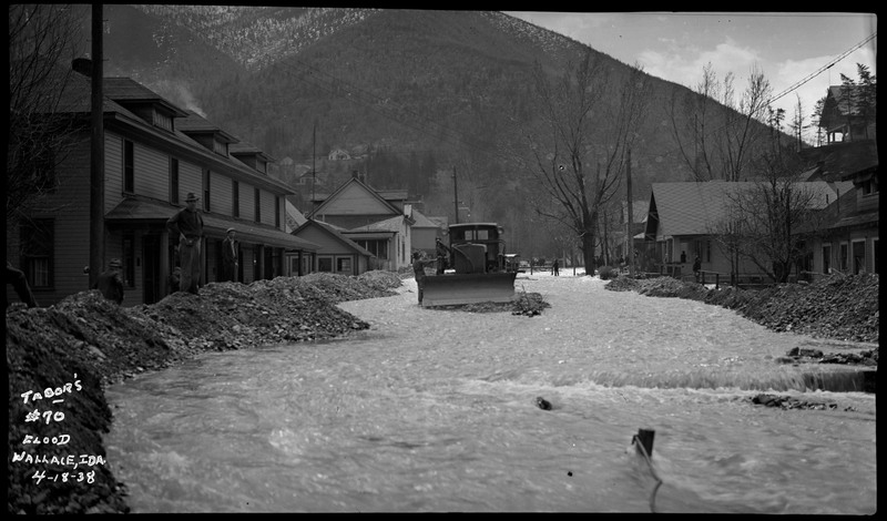 Street view of Wallace, Idaho during a flood. A tractor is stuck in some debris as water flows by. There are standing buildings behind piles of rocks to prevent flooding. Several men can be seen, some standing in the distance and others stand closer to the camera. 
